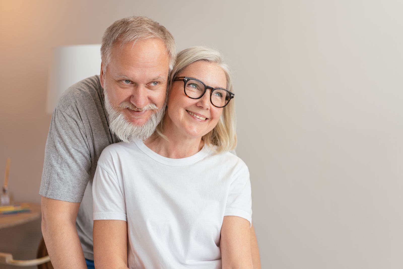 Smiling senior couple sharing a loving embrace indoors, both wearing eyeglasses.